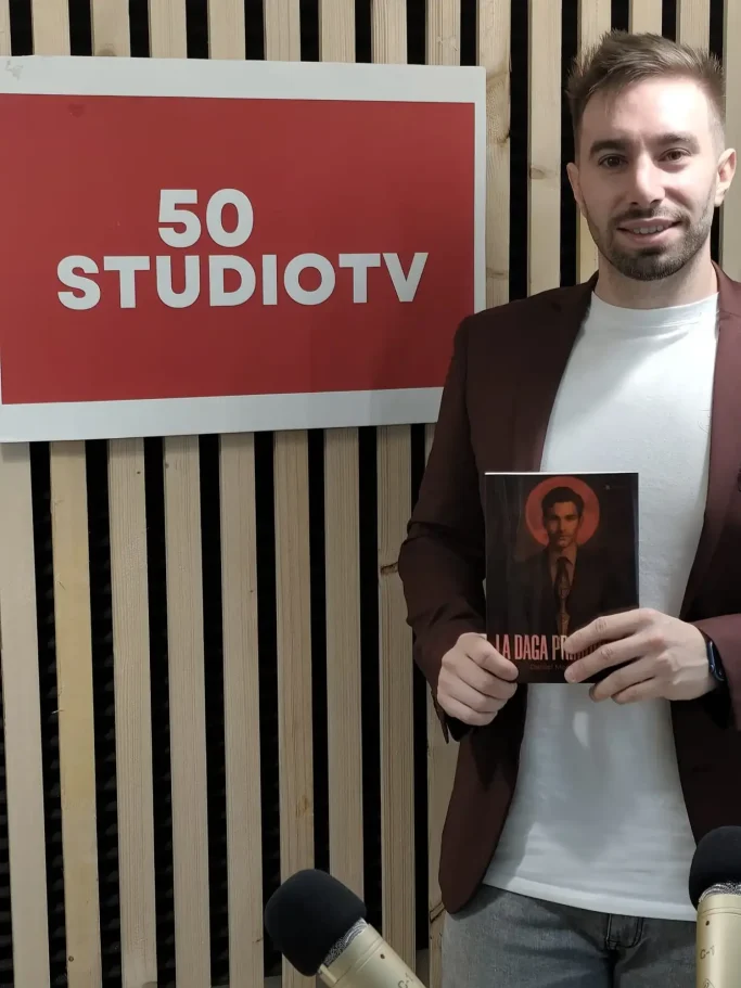 Hombre sosteniendo un libro frente a un cartel de 50 STUDIO TV.