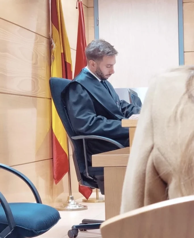Dani en una sala de tribunal, con banderas de España de fondo, revisando documentos.