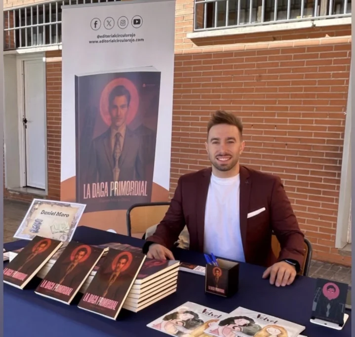 Hombre sonriente sentado detrás de una mesa con libros y material promocional.